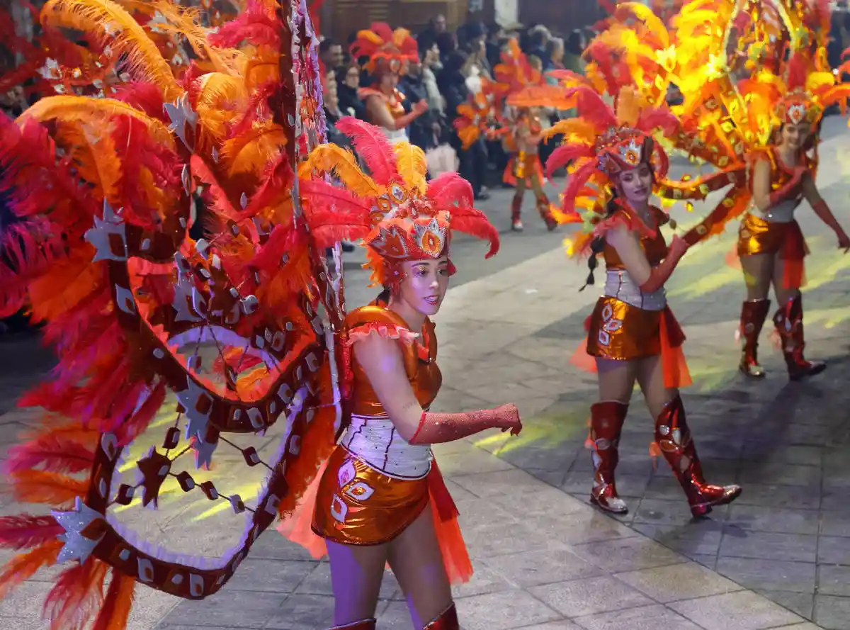 Comparsas y Color en el Carnaval de Poio Comparsas con vistosos disfraces de plumas rojas y naranjas desfilando en el Carnaval de Poio, rodeadas de público y luces festivas.